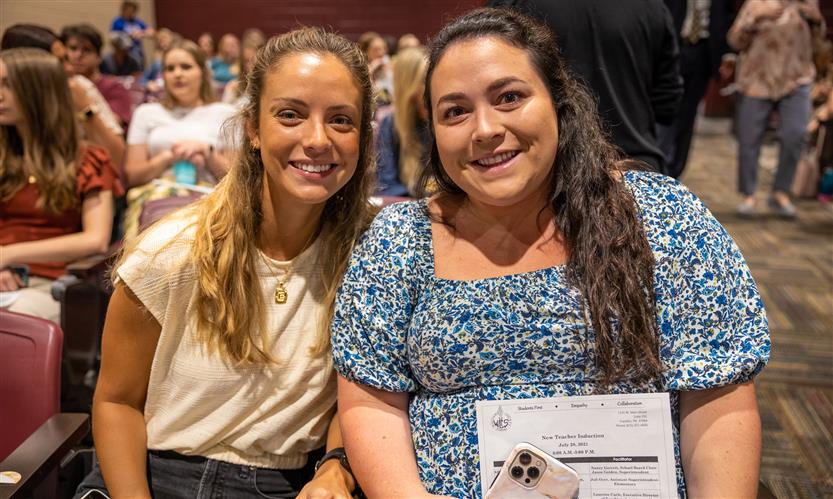 Two women in an auditorium lean together and smile